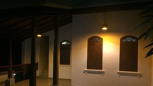 L’extérieur d’un bâtiment de nuit, avec deux fenêtres illuminées, une porte en bois et un escalier menant à un étage supérieur.