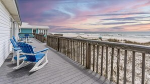 A wooden deck with blue Adirondack chairs, a beach view, and a building with a white facade.