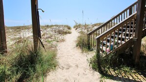 A sandy path leading to a wooden staircase with railings, surrounded by vegetation.