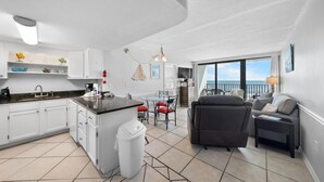 A modern kitchen with white cabinets, a dining area with a glass table, and a living room with a sofa and a balcony with a view. - Island Sunrise 362-The Beach is Where You Need to Be! (Gulf Shores)