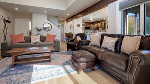 Un salón espacioso con un amplio sofá de cuero marrón, un sofá gris con cojines naranja y una mesa de centro de madera. Una alfombra estampada cubre el suelo, y hay una escalera que conduce a un nivel superior.