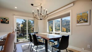 Un comedor con una gran mesa de madera, sillas negras y una araña. Se observan amplias ventanas que ofrecen vistas a un paisaje montañoso.