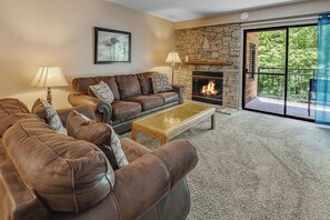 A living room with a stone fireplace, a brown leather sofa, a wooden coffee table, and a large sliding glass door leading to a balcony. - two bedroom condo sleeps 6 persons (Gatlinburg)