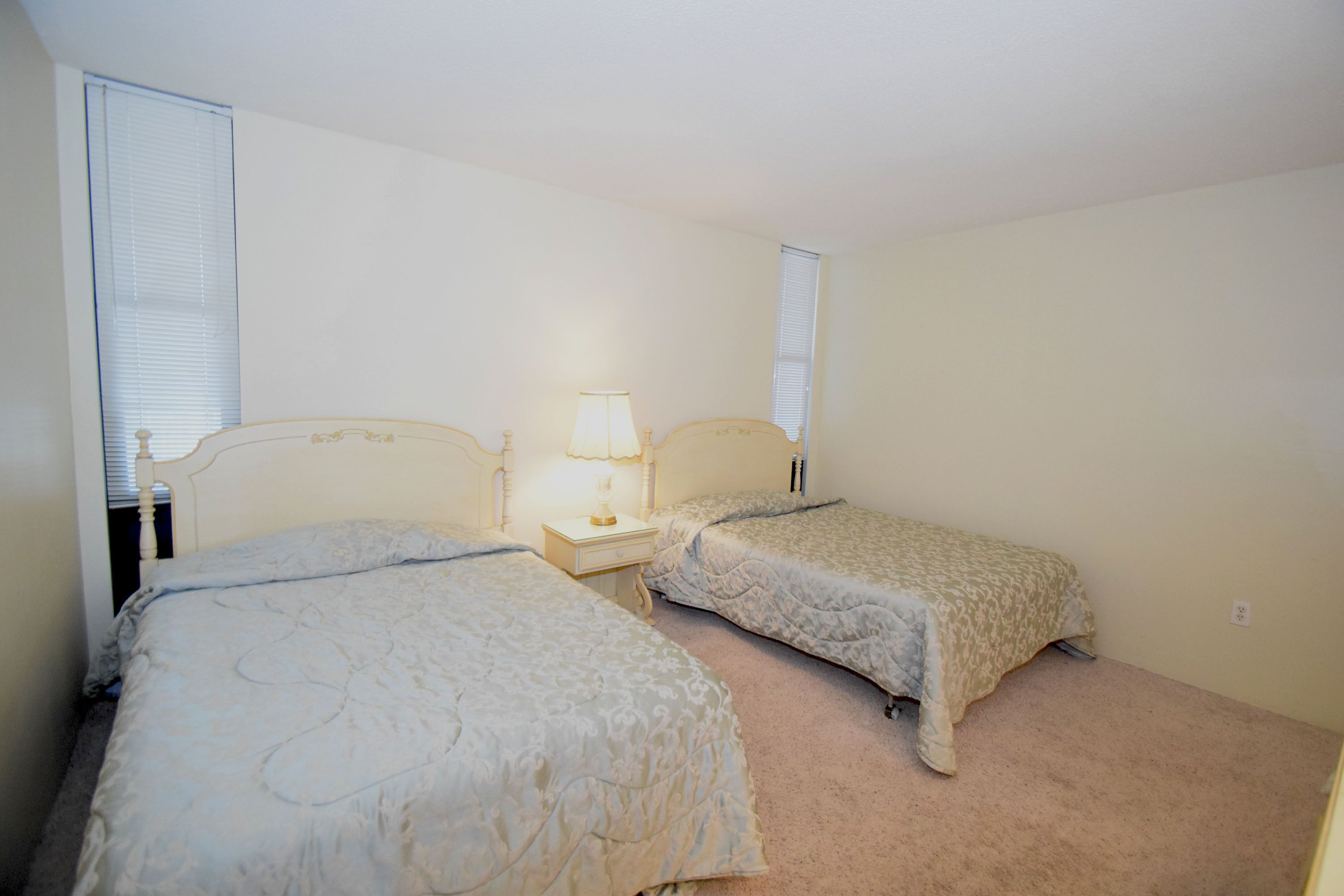 A bedroom with two beds, a nightstand with a lamp, and a window with blinds.