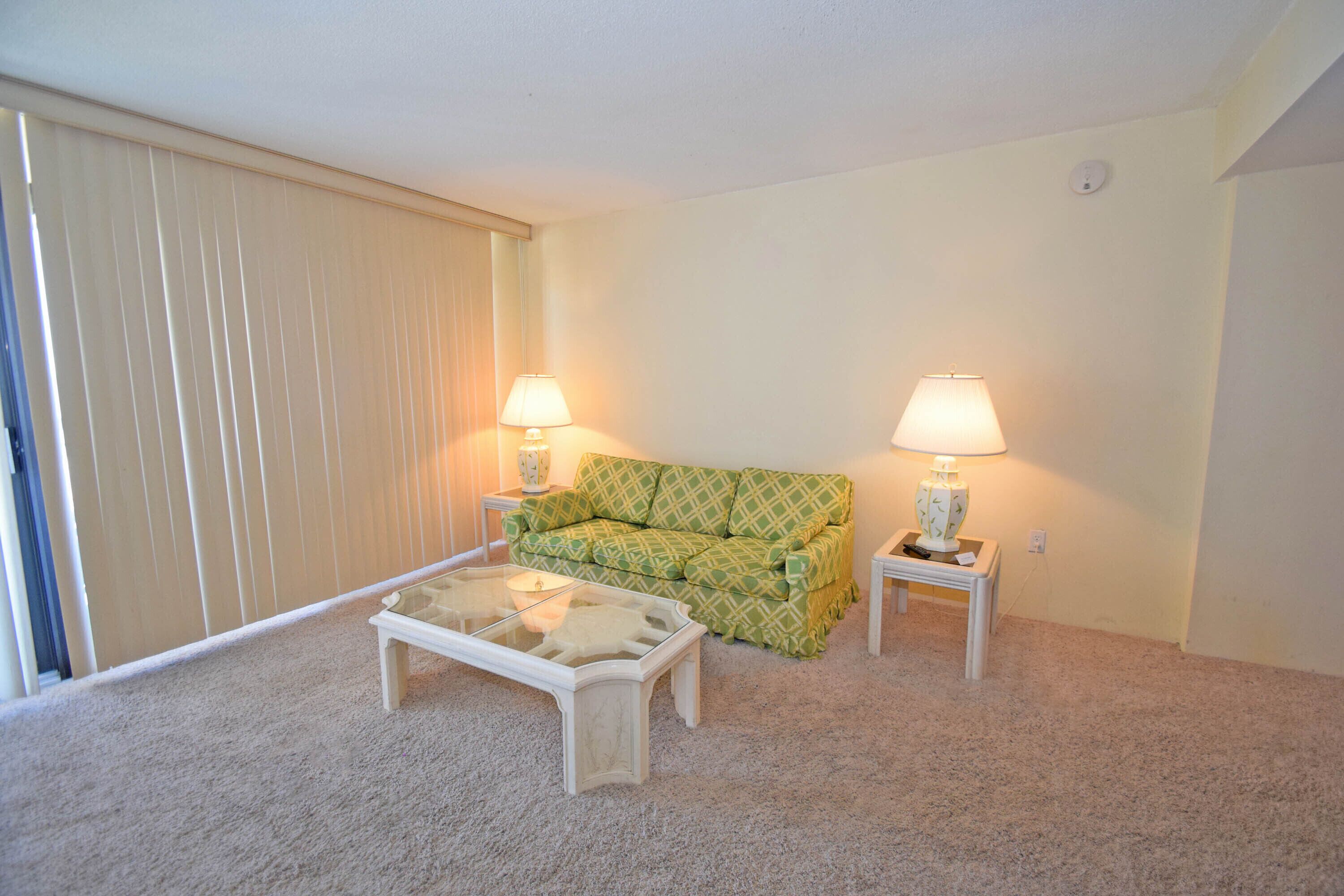 Ein Wohnzimmer mit einem grün gemusterten Sofa, zwei weißen Nachttischen mit Lampen, einem Glastisch und einem beigen Teppich.