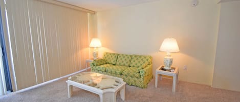 Ein Wohnzimmer mit einem grün gemusterten Sofa, zwei weißen Nachttischen mit Lampen, einem Glastisch und einem beigen Teppich.