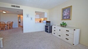 A living room with a dining area, a kitchen in the background, and a white dresser with drawers. - Fountainhead 702-Oceanfront 117th St, WIFI, Pool, Elev, W/D, AC (Ocean City)