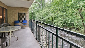 Ein Balkon mit Betonboden, zwei blaue Stühle, einem Glastisch und Blick auf einen Fluss mit Steinen und Bäumen.