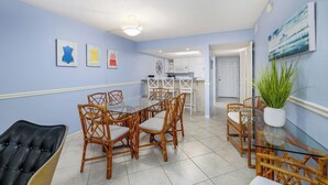 A dining area with a glass table and wicker chairs, a kitchen in the background, and a potted plant on the right. - Slice Of Paradise (Sanibel)