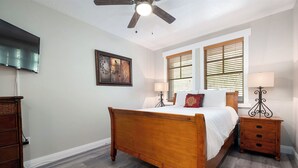 A bedroom with a wooden bed, two bedside tables with lamps, a ceiling fan, a TV mounted on the wall, and a window with blinds. - Cozy Hotel Suite-Block from Main Street and Lift I Managed by Park City Lodging (PARK CITY)