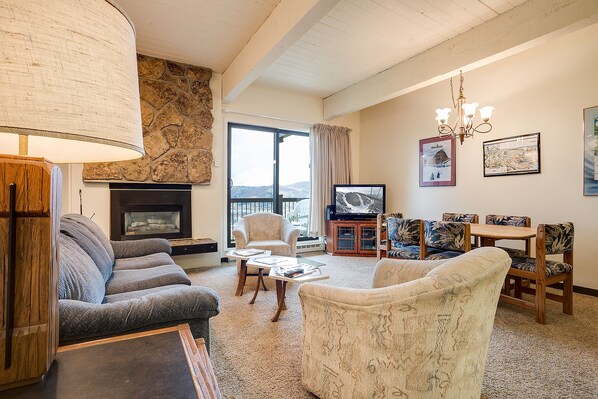 A cozy living room with a stone fireplace, comfortable seating, a television, and a view of the outdoors.