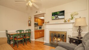 A living room with a fireplace, a television mounted above it, and a dining area with wooden cabinets and chairs.