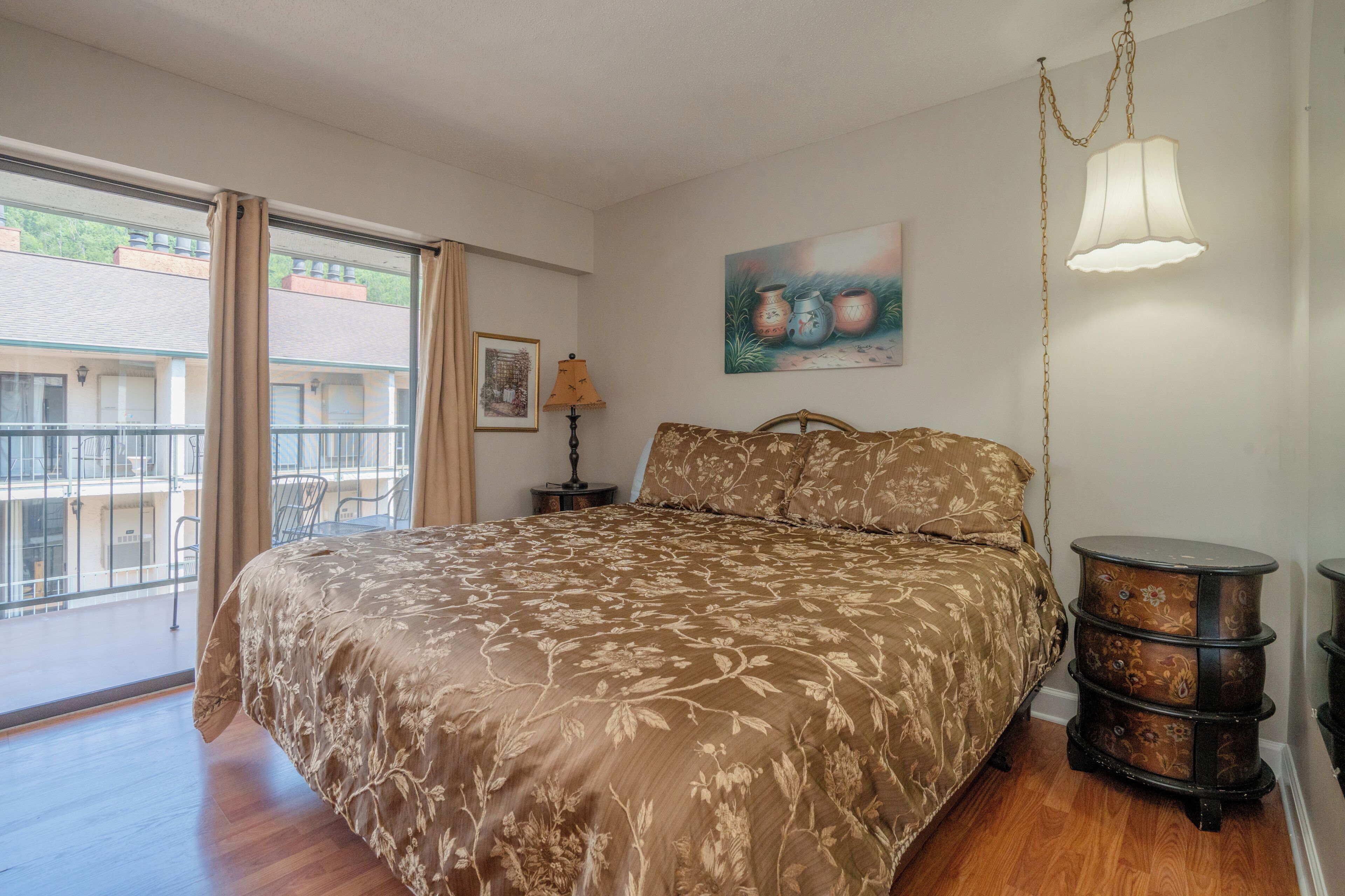 A bedroom with a large bed, a side table, a lamp, a painting, and a view of the outside.