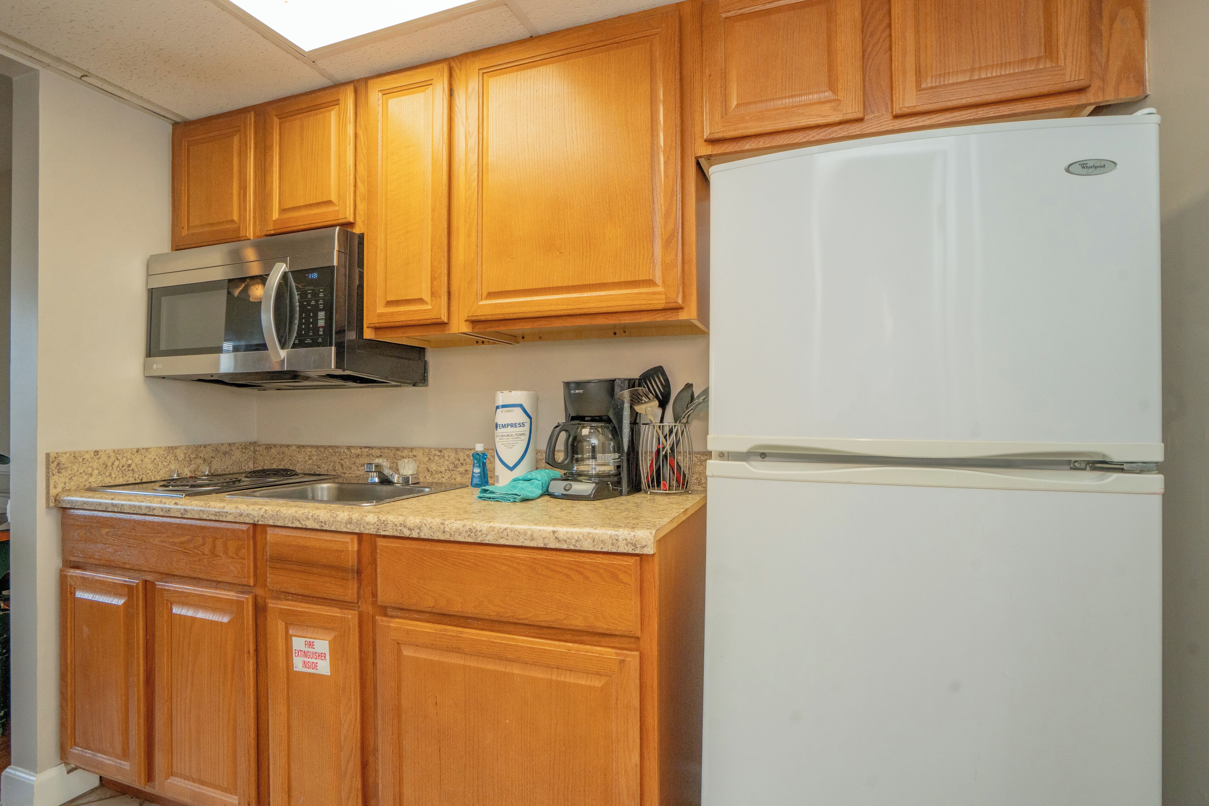 Una cocina con gabinetes de madera, un microondas de acero inoxidable, un refrigerador blanco, una cafetera y una botella de detergente para platos.
