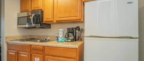 Una cocina con gabinetes de madera, un microondas de acero inoxidable, un refrigerador blanco, una cafetera y una botella de detergente para platos.