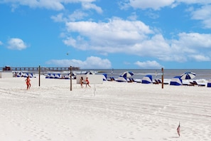 A beach with umbrellas, a volleyball net, and a pier in the background.