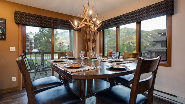 Un espace repas avec une grande table dressée pour six personnes, entourée de chaises, agrémenté d’un lustre et offrant une vue sur un paysage montagneux grâce à de grandes fenêtres.