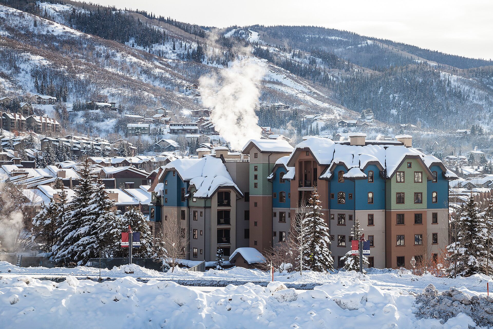 Un bâtiment enneigé, avec une cheminée d’où s’échappe de la fumée, entouré d’arbres chargés de neige et avec une chaîne de montagnes en arrière-plan.