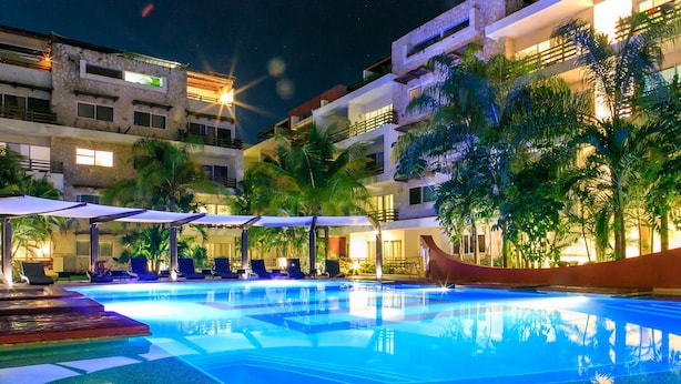 A swimming pool with lounge chairs and umbrellas, surrounded by palm trees and a multi-story building with balconies. - Incredible- Fully funrished & equipped+ pool view - 1 block from 5th (Playa del Carmen)