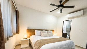 A hotel room with a bed, nightstands, a ceiling fan, a mirror, and a bathroom visible through a glass door. - Incredible- Fully funrished & equipped+ pool view - 1 block from 5th (Playa del Carmen)
