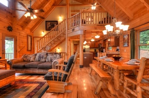 A spacious cabin with a wooden interior, a dining area, a living room with a sofa and chairs, and a staircase leading to an upper level. - 3BR log cabin, Hot Tub, sleeps 9, exceptional view + free railroad tix (Bryson City)
