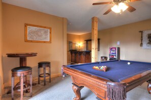 A pool table with a wooden frame, two stools, a ceiling fan, and a neon arcade machine. - 3BR log cabin, Hot Tub, sleeps 9, exceptional view + free railroad tix (Bryson City)