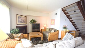 A living room with a sofa, coffee table, and a television. There is a staircase to the right and a plant on the wall.
