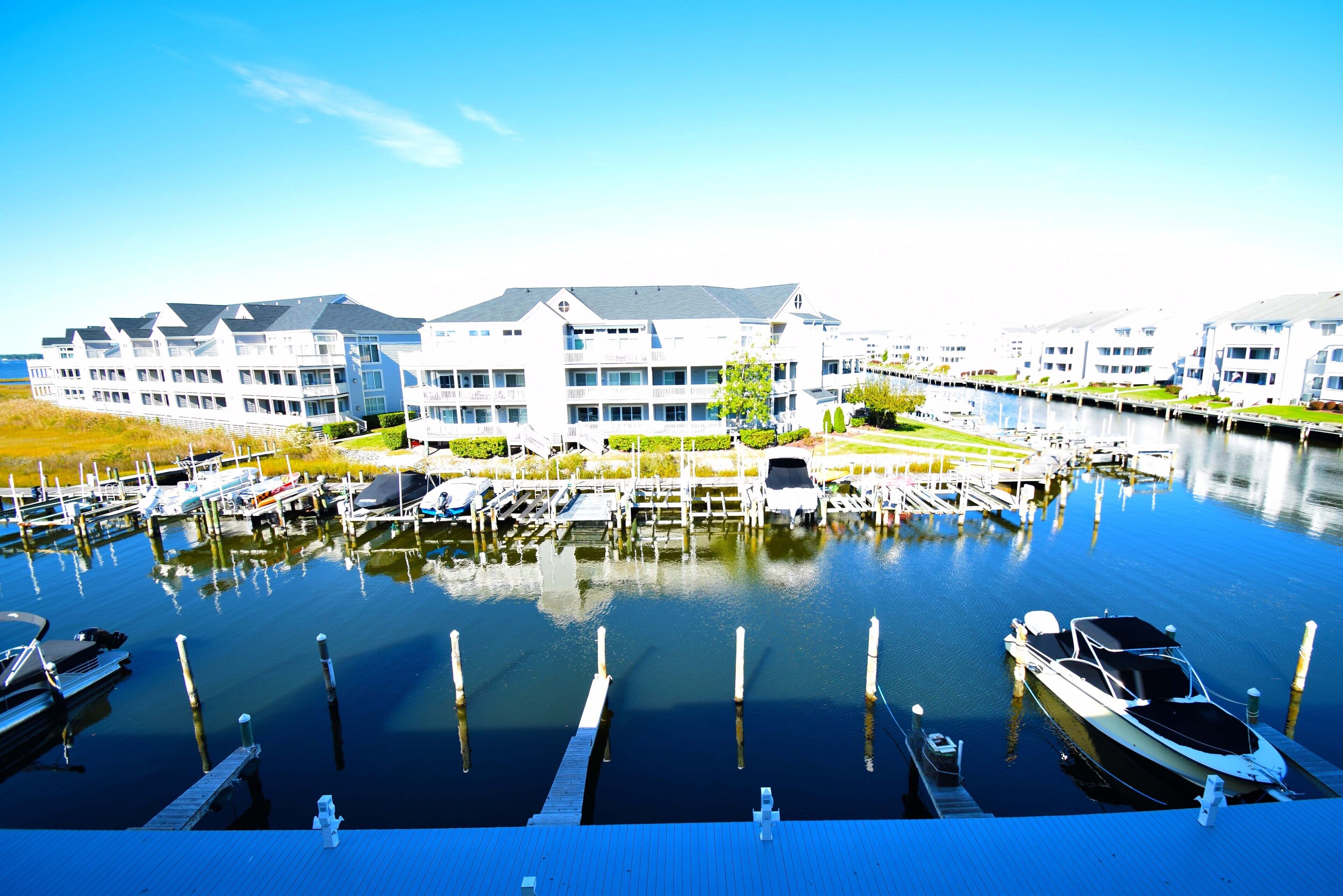 A marina with several boats docked, a dock in the foreground, and a row of modern buildings with balconies in the background.