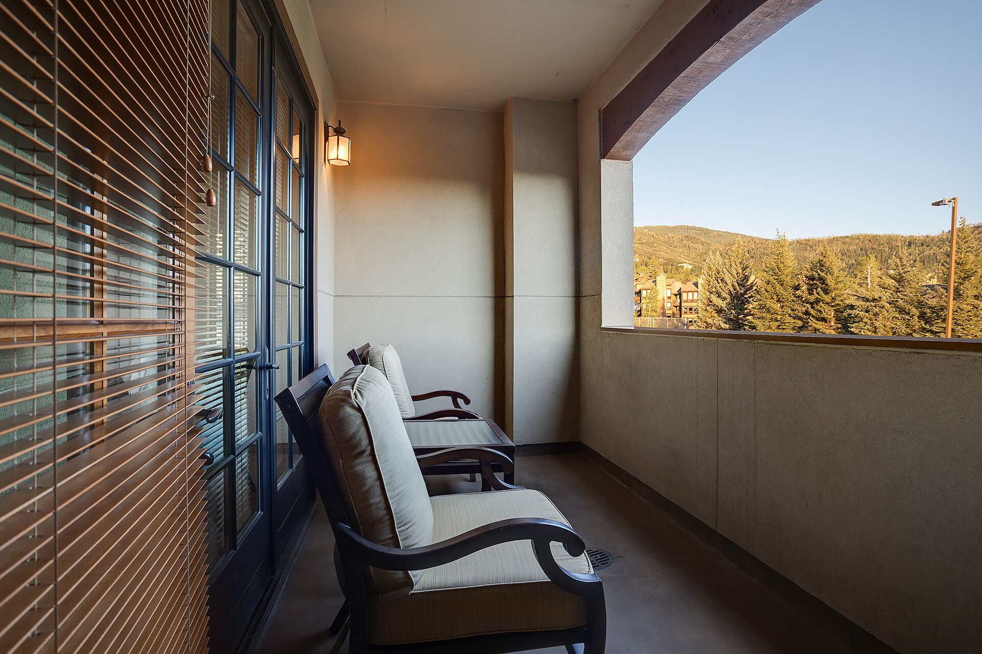 A balcony with two chairs, a window with blinds, and a view of a forested area.