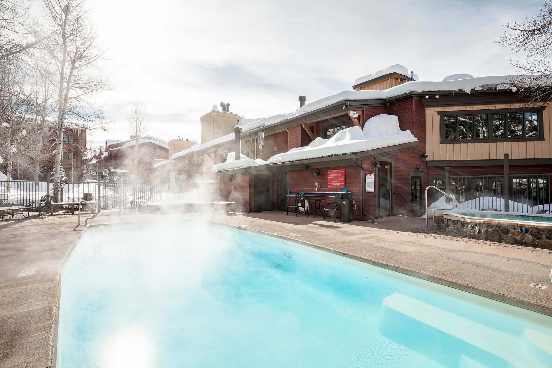 A pool area with a hot tub, benches, and a building with a snow-covered roof.