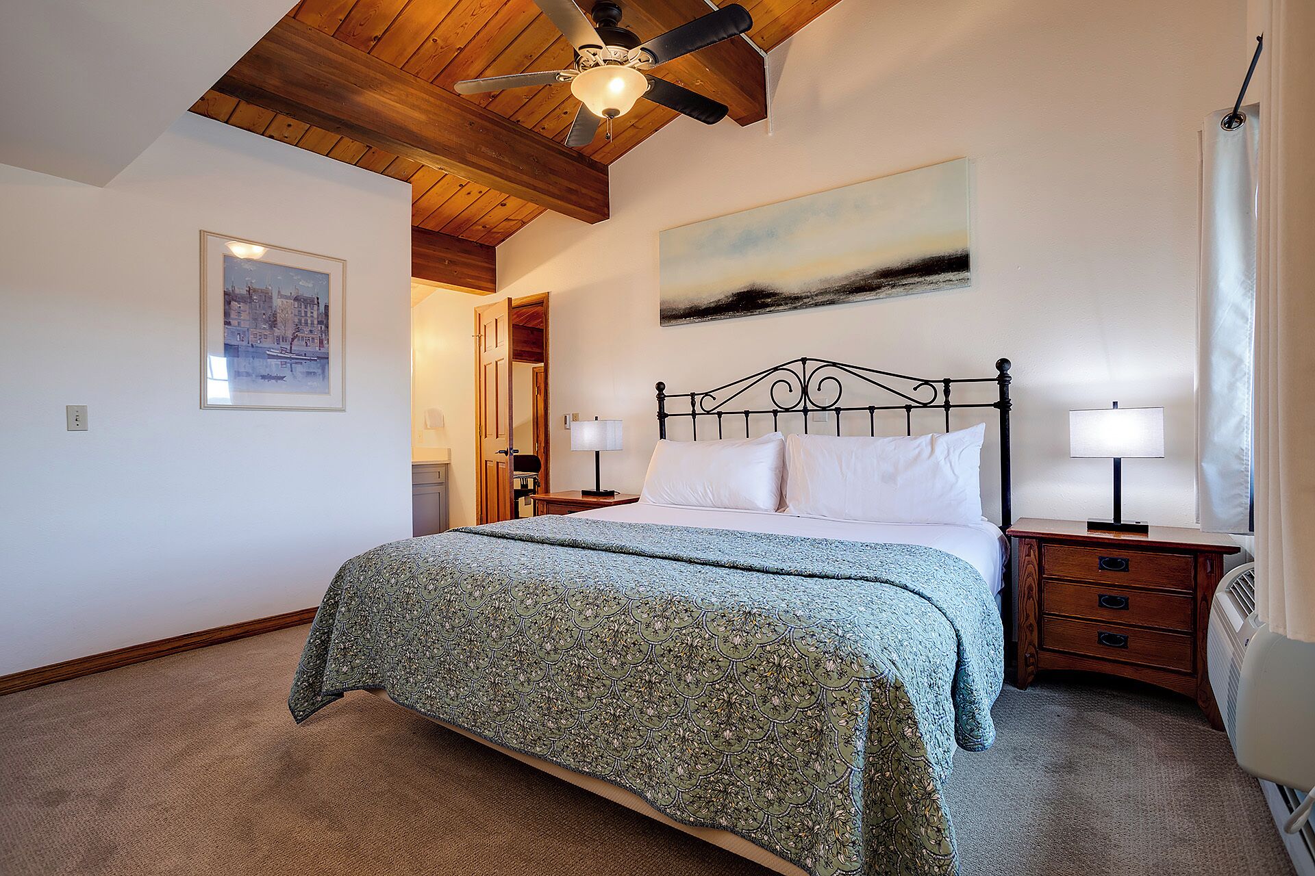 A bedroom with a bed, two bedside tables, a ceiling fan, a painting, and a door leading to another room.
