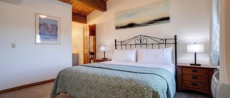 A bedroom with a bed, two bedside tables, a ceiling fan, a painting, and a door leading to another room.
