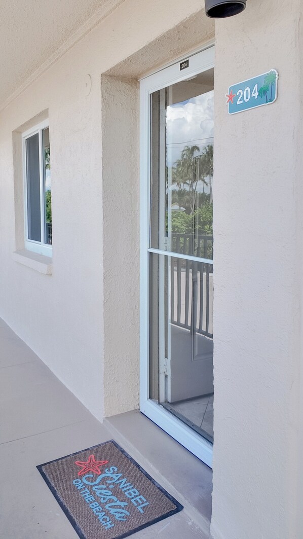 A building entrance with a number sign '204', a doormat with 'Sanibel Beach' text, and a window with a view of palm trees. - Taste Of Paradise (Sanibel)