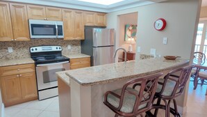 A kitchen with wooden cabinets, a stainless steel refrigerator, a microwave, a stove, and a granite countertop. There are two bar stools with cushioned seats. - Taste Of Paradise (Sanibel)