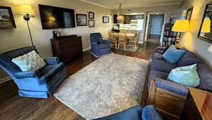 A living room with a blue sofa, a blue armchair, a dining table with chairs, a flat-screen TV, and a kitchen area in the background. - Cozy, yet spacious 2 bedroom oceanfront condo with free WiFi and a stunning view of the ocean located midtown just a few steps to the beach! (Ocean City)