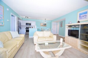 A living room with two beige sofas, a glass coffee table, a TV, and a kitchen in the background.