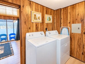 A laundry room with two white washing machines, wooden walls, a window with a view, and a blue chair.