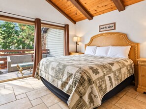 A bedroom with a large bed, a nightstand, a lamp, a wooden headboard, and a view of the outdoors.