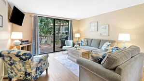 Ein Wohnzimmer mit Sofa, Sesseln und Couchtisch. Ein Fernseher ist an der Wand befestigt und es gibt ein Fenster mit Vorhängen.