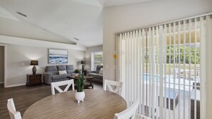 Un coin repas avec une table ronde, des chaises blanches et une plante en pot. On aperçoit un canapé gris et une lampe à l’arrière-plan.