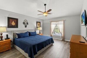 A bedroom with a bed, nightstands, a ceiling fan, a window with curtains, and artwork on the walls.