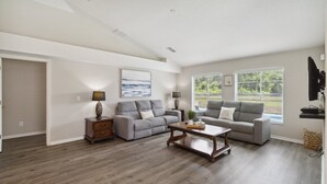 Un salon spacieux doté d’un parquet, de deux canapés gris, d’une table basse en bois et d’une grande fenêtre avec des stores.