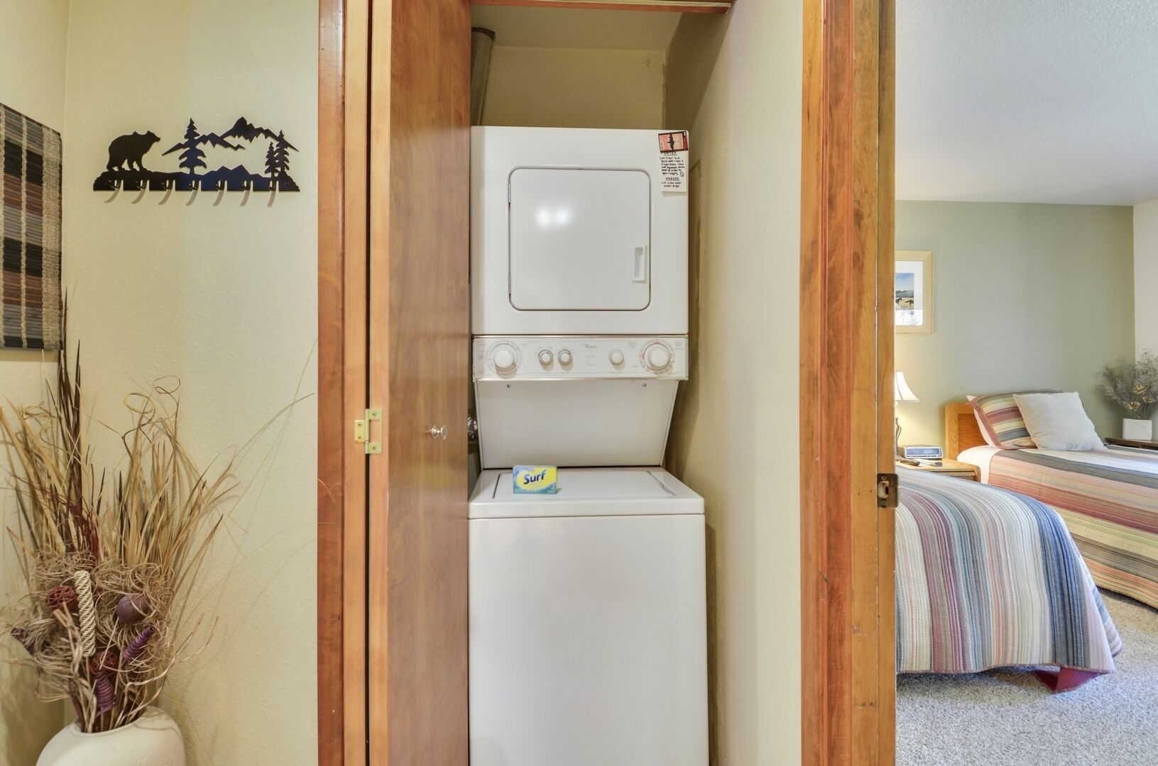 A compact laundry room with a washing machine and dryer, a bed with a striped bedspread, and a wall decoration of a bear and trees.