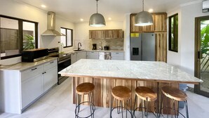 Een moderne keuken met een groot eiland, houten barkrukken, roestvrijstalen apparatuur en hangende lampen.