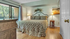 A bedroom with a large bed, two bedside lamps, a decorative headboard, a small table, and a view through a window. - two bedroom condo sleeps 4 persons (Gatlinburg)