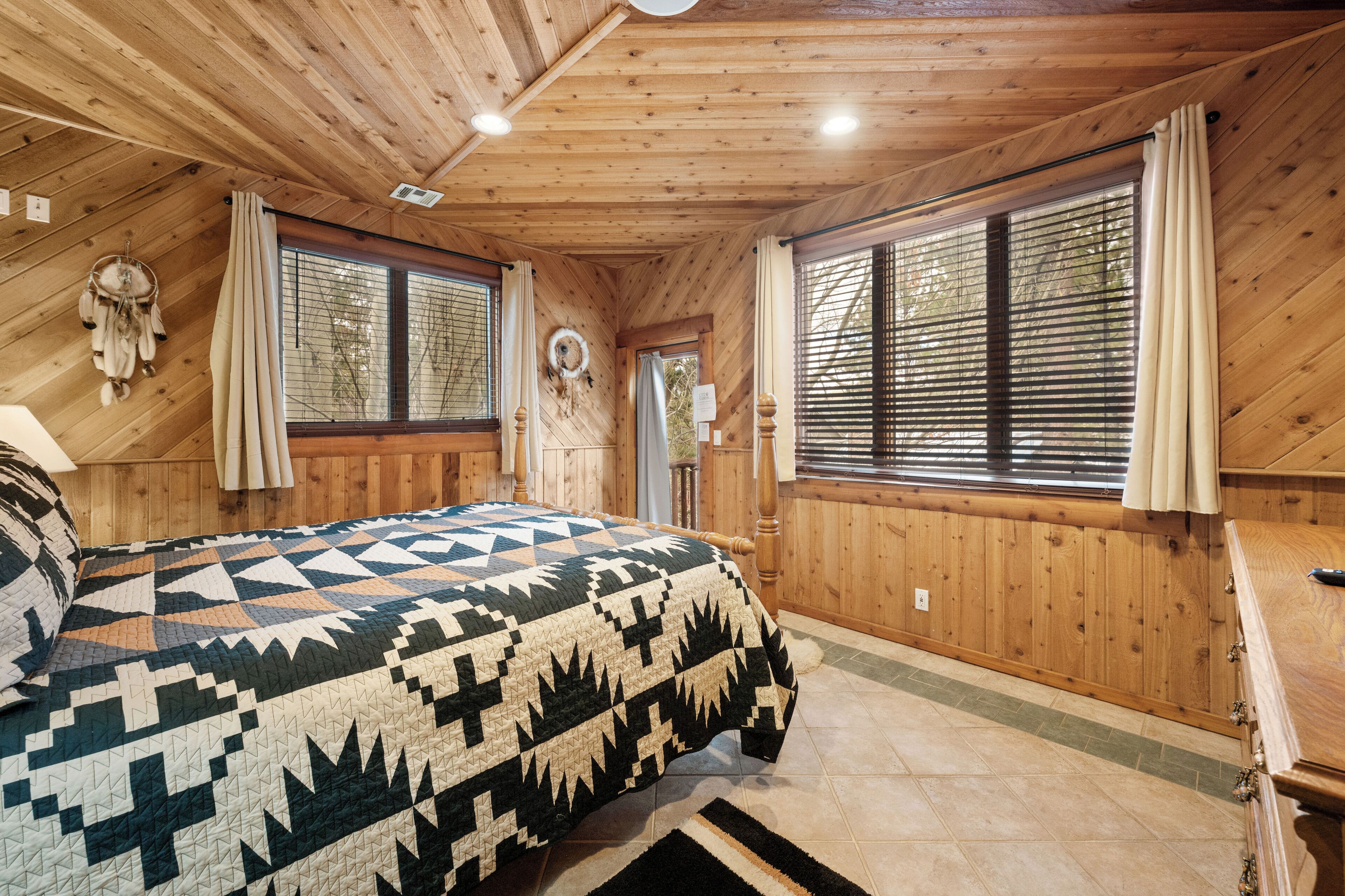 Een slaapkamer in een houten blokhut met een bed, ramen en een dromenvanger aan de muur.