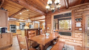 A wooden cabin interior with a dining area, kitchen, and large windows.
