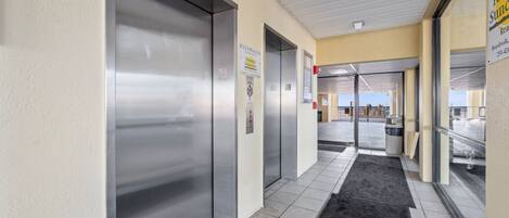 Un passadís amb ascensors, una catifa negra i una porta de vidre que dona a zona exterior.