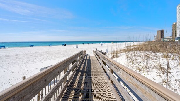 A wooden boardwalk leading to a sandy beach with clear blue water and a few buildings in the background.