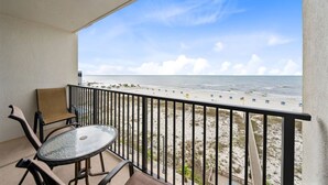 A balcony with a glass table and chairs, overlooking a beach with umbrellas and people.